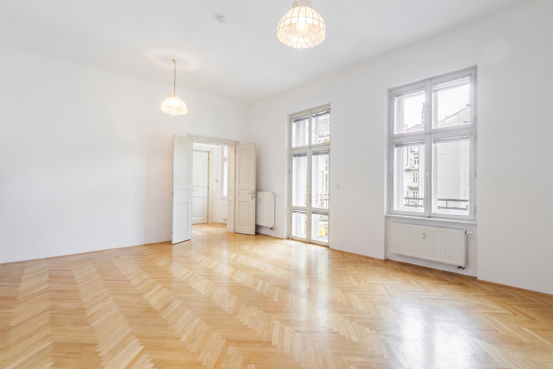Bright, empty room with white walls, herringbone wood floors, two pendant lights, and large windows overlooking a city.