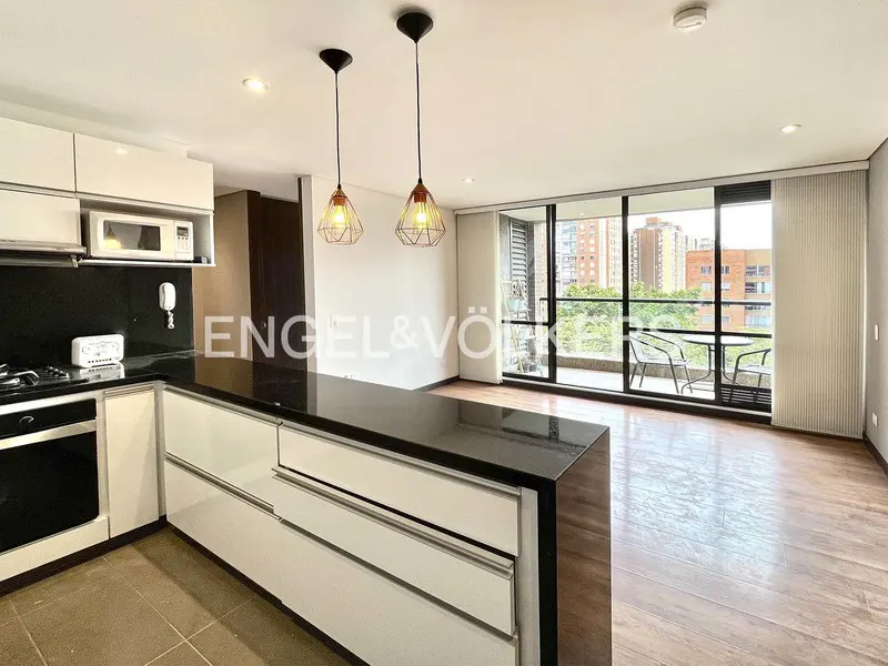 A modern kitchen with white cabinets, black countertops, and wood floors leads to a balcony with city views. Two pendant lights hang above the island.