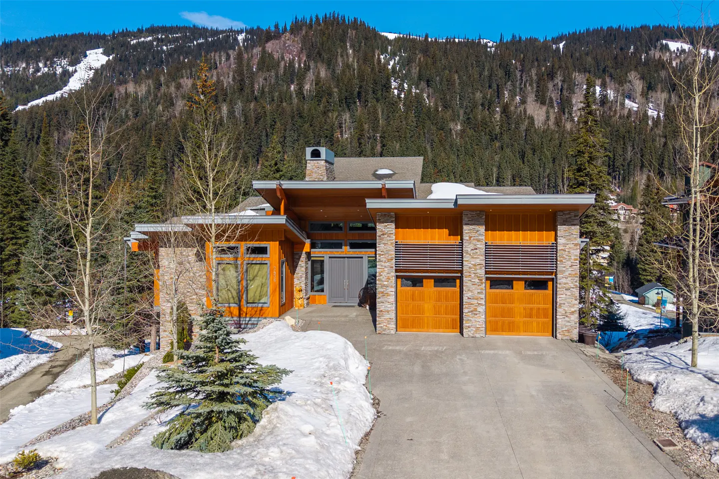 Modern mountain home with wood and stone exterior, gray roof, and two-car garage. Snow on the ground and forested mountain in the background.
