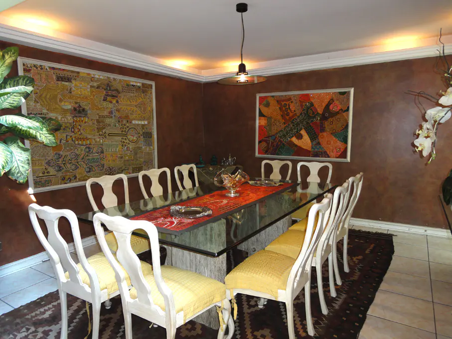 Dining room with brown walls, a glass-topped table, and white chairs with yellow cushions. Two framed art pieces hang on the walls.