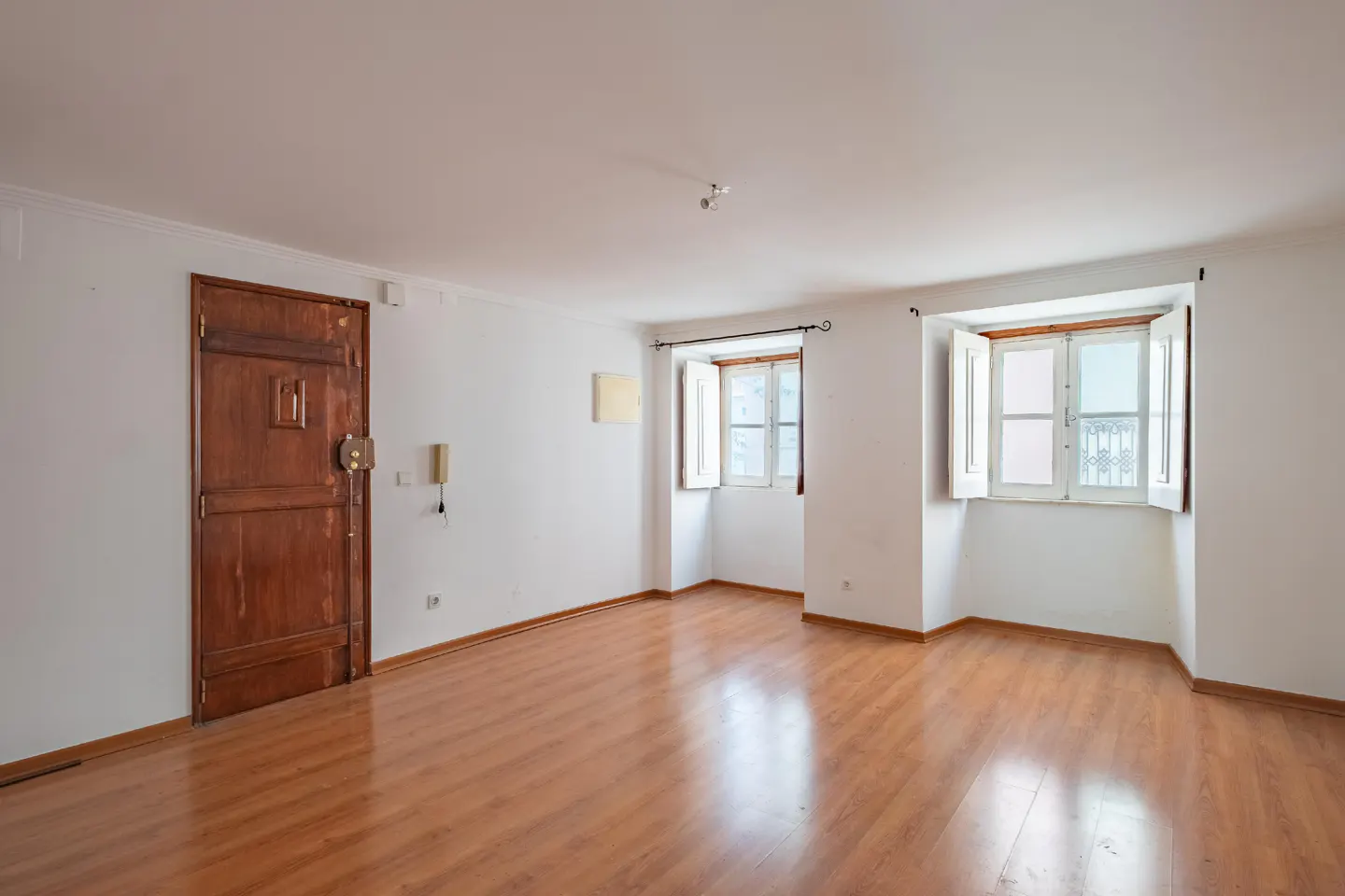 Empty room with wood floors, white walls, and two windows with white shutters. A brown door is on the left.