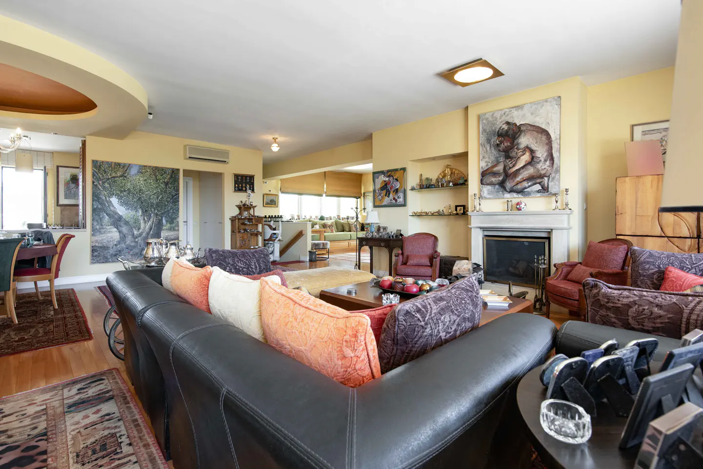 A living room with a black leather sofa, colorful pillows, and artwork on the yellow walls. A fireplace and various furniture pieces are visible.