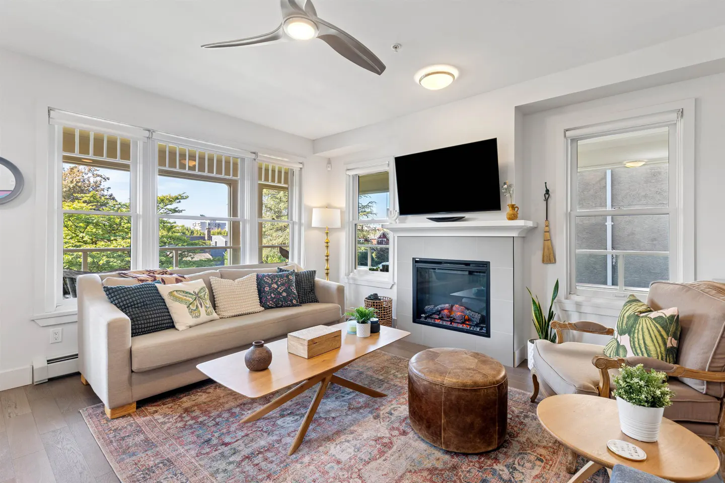 Bright living room with beige sofa, wood coffee table, fireplace, and large windows overlooking green trees.