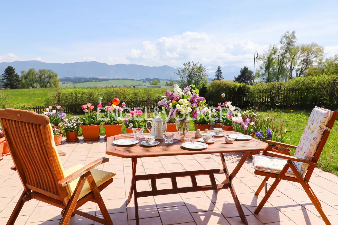 Outdoor patio with wooden table set for tea, surrounded by colorful flowers and greenery, with mountains in the background.