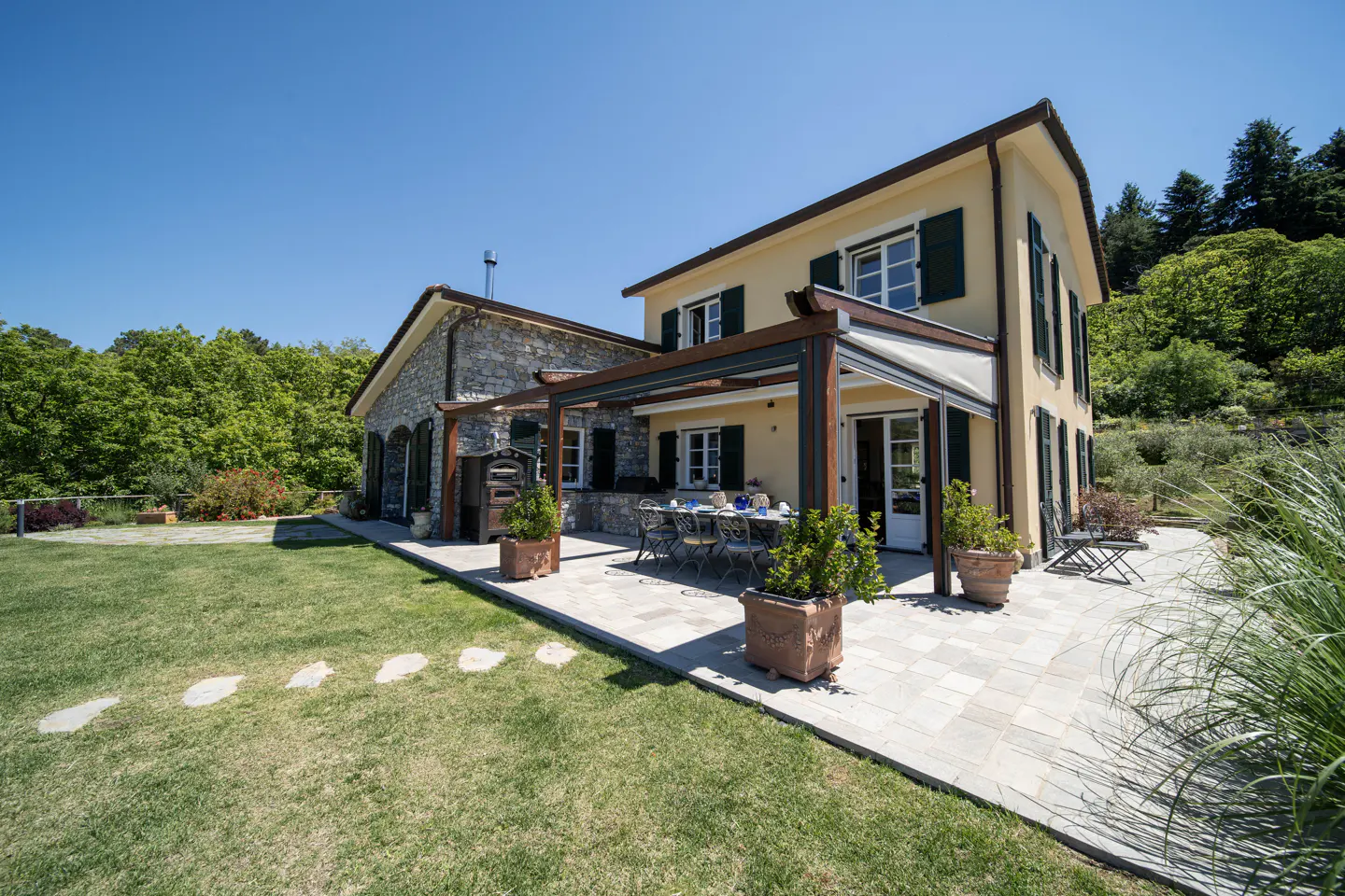 Exterior view of a two-story house with a stone patio, table, and green lawn under a clear blue sky.