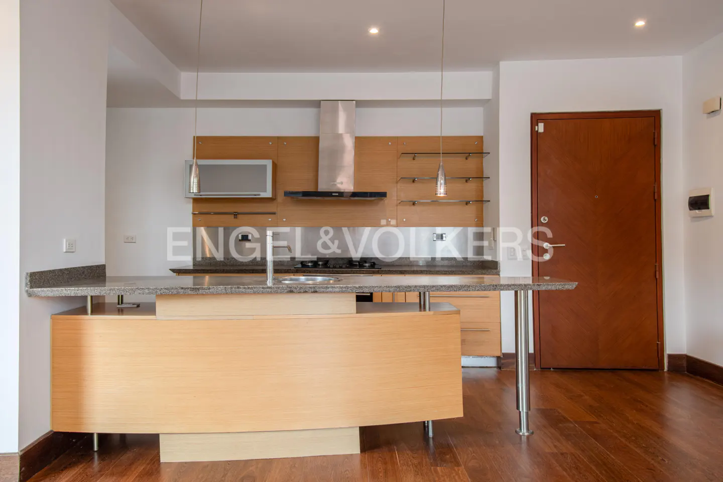 A modern kitchen with light wood cabinets, stainless steel appliances, and a granite countertop island. Hardwood floors and a brown door are visible.