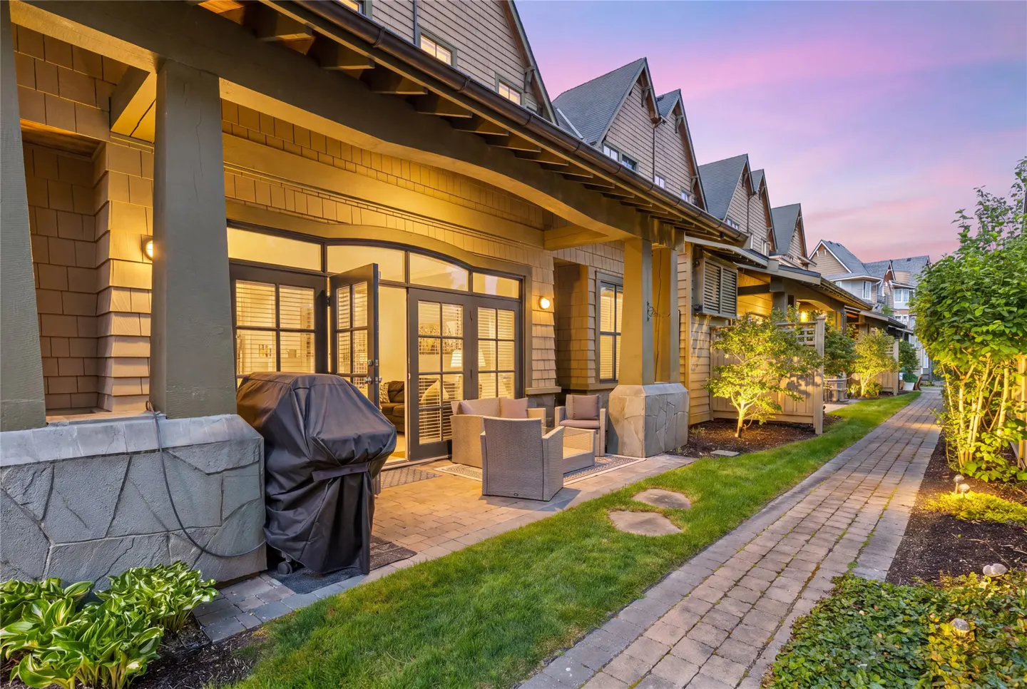 Exterior view of townhouses with patios, a grill, and a brick walkway at dusk.