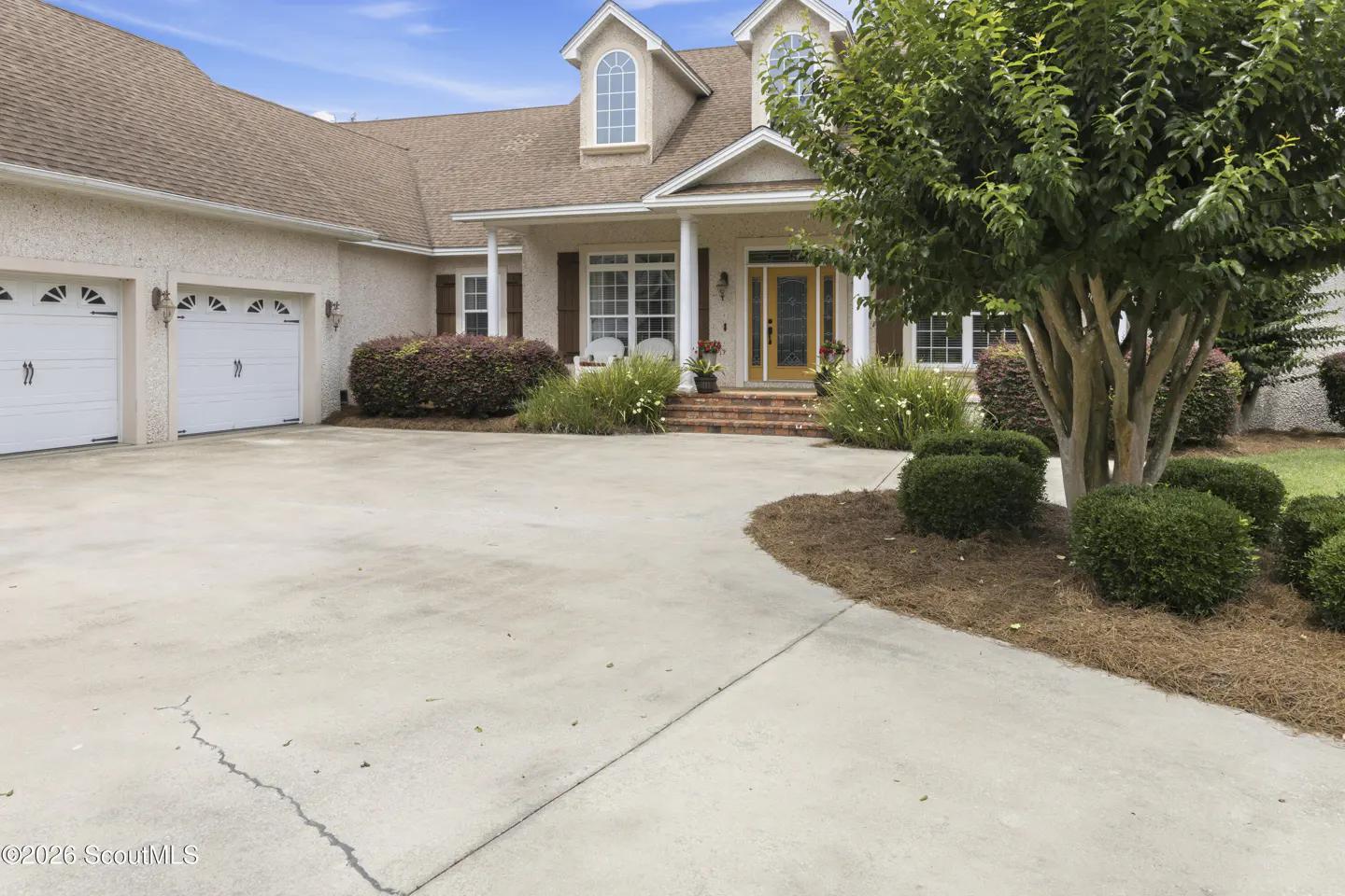 Beige stucco house with a brown roof, white garage doors, and a yellow front door. A large tree is on the right.