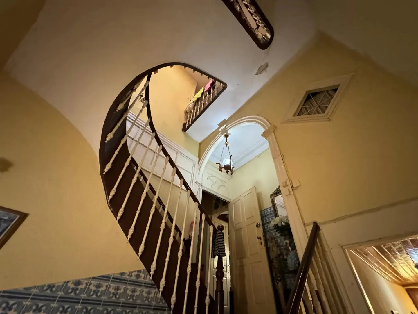Interior view of a home's foyer with a curved staircase, brown wood banister, white spindles, and yellow walls. A doorway leads to another room with a chandelier.