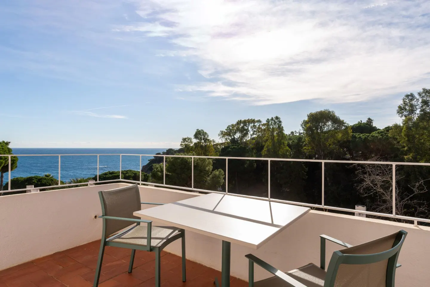Balcony with a white table and two chairs overlooking the ocean and trees on a sunny day.