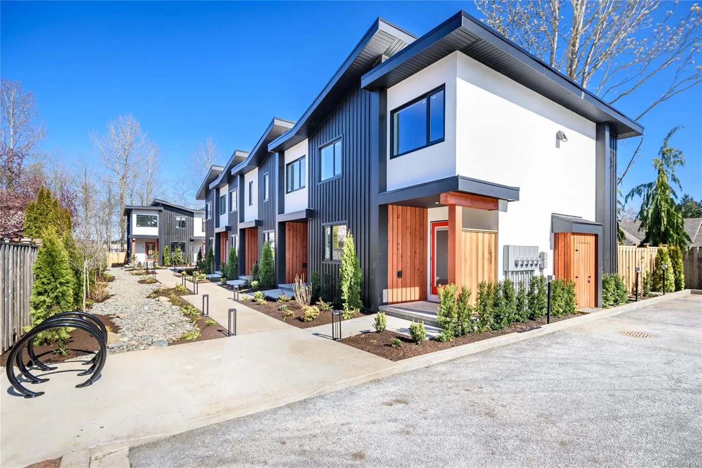 Row of modern townhouses with black, white, and wood exteriors under a blue sky. A concrete walkway leads to the red front doors.
