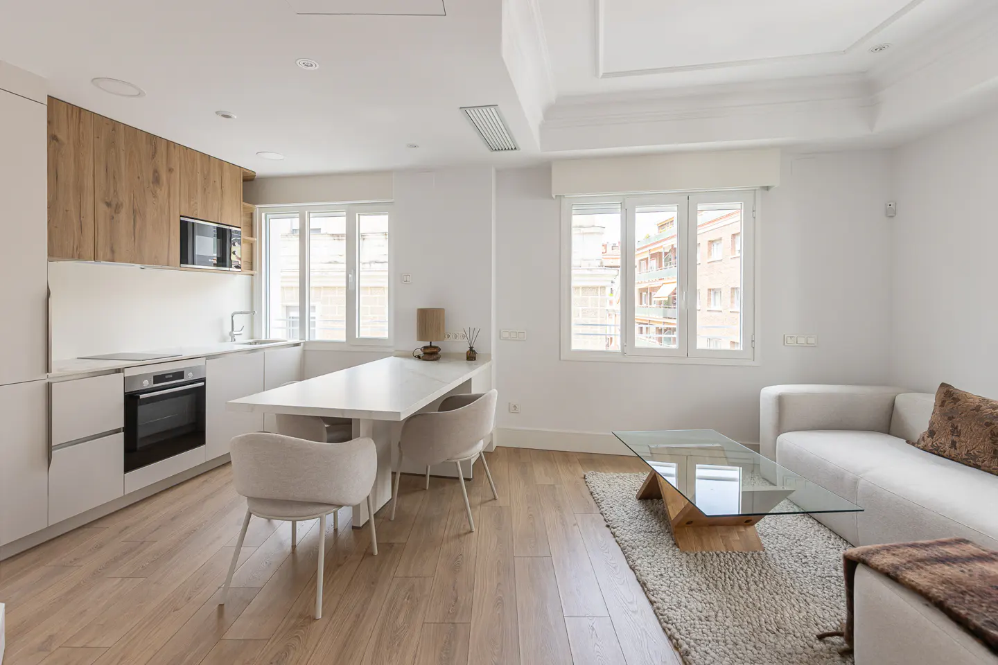 Bright, modern apartment with white walls, wood floors, and large windows. Kitchen with white cabinets and island, living area with sofa and glass table.