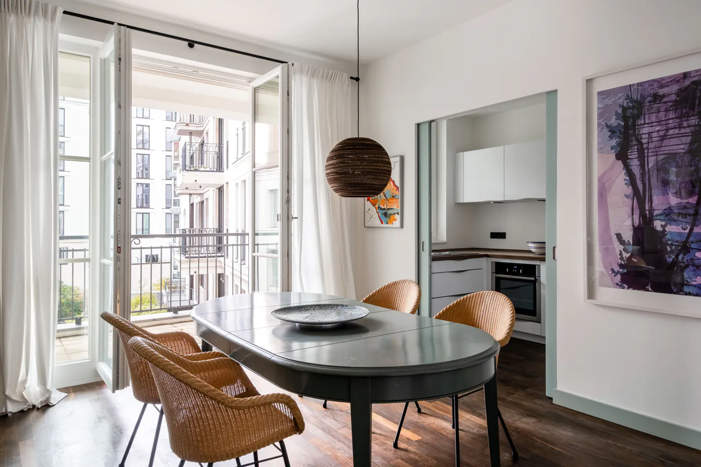 Bright dining room with a gray oval table, wicker chairs, and open French doors to a balcony. A modern kitchen is visible in the background.
