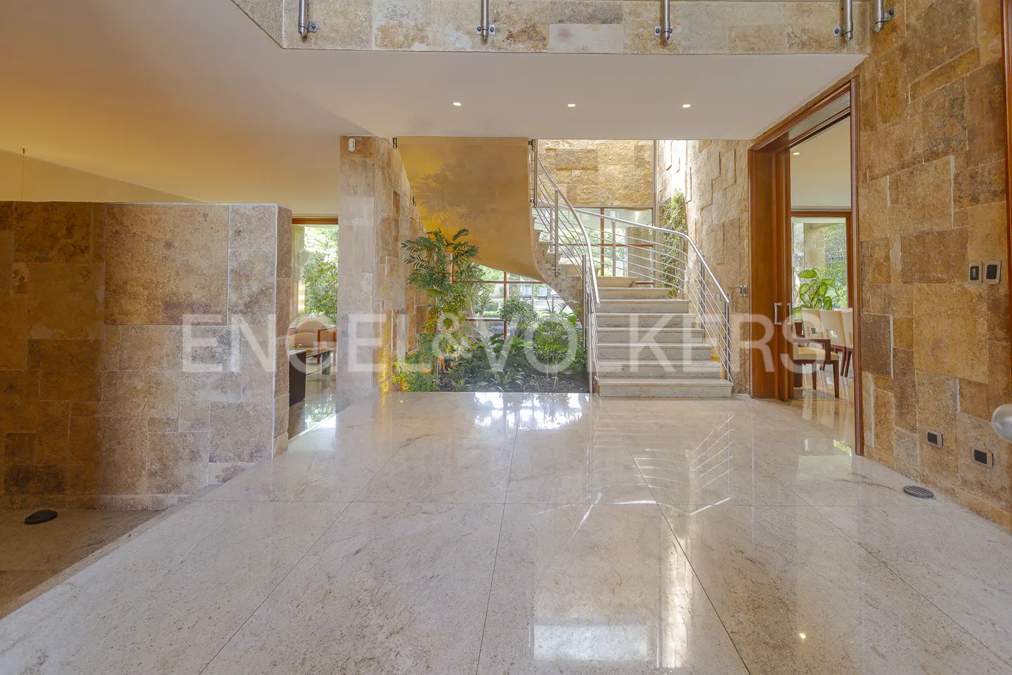 A grand foyer with stone walls, a marble floor, and a staircase with stainless steel railings. Green plants add a touch of nature.