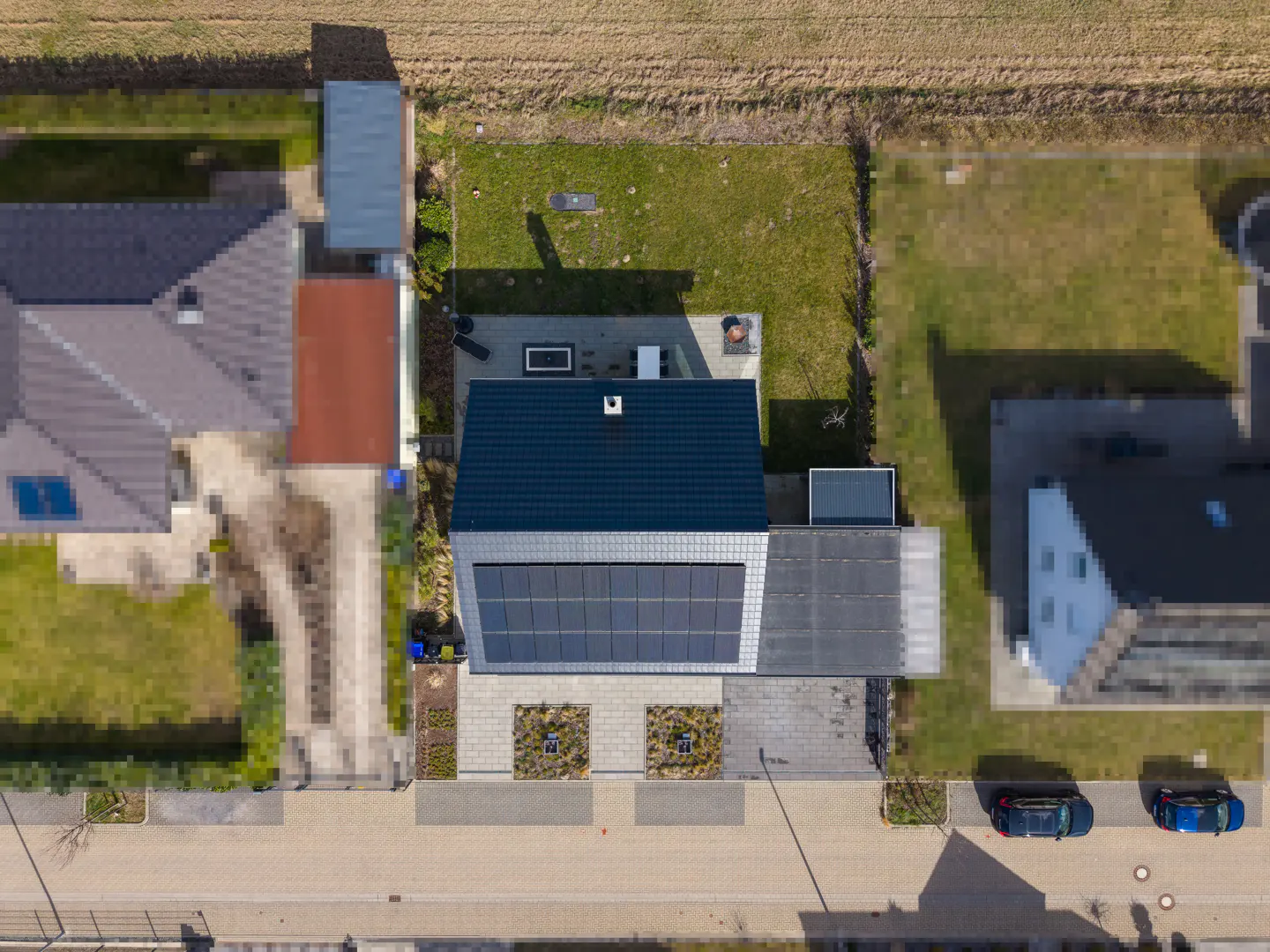 Aerial view of a modern house with solar panels, a blue roof, and a green lawn, situated on a street with parked cars.