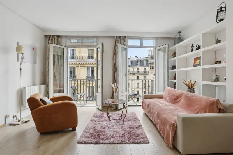 Bright living room with open balcony doors revealing Parisian buildings. A tan armchair and pink sofa sit on a wood floor with a pink rug.