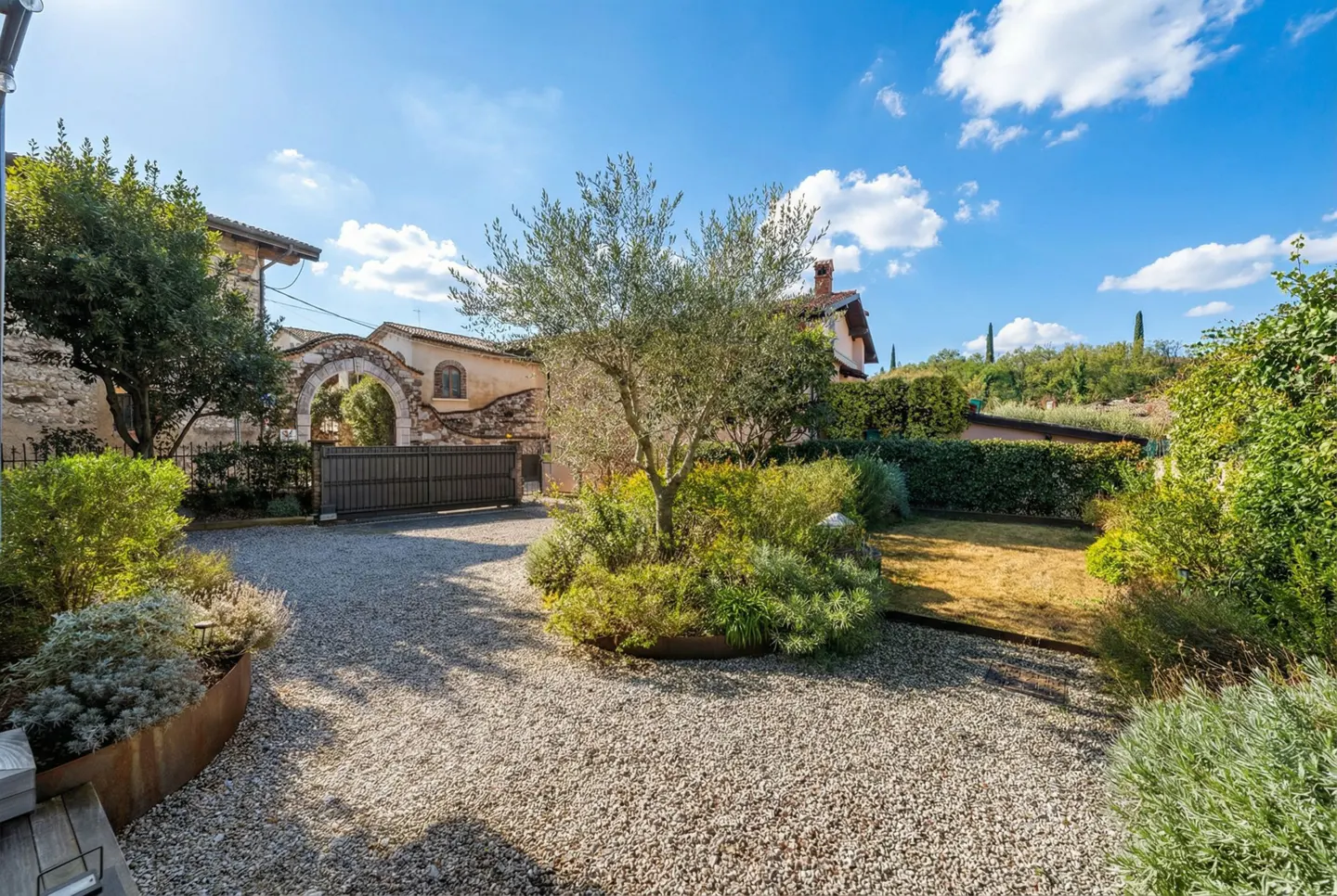 Exterior view of a gravel driveway leading to a stone archway with a metal gate, surrounded by greenery and a blue sky.