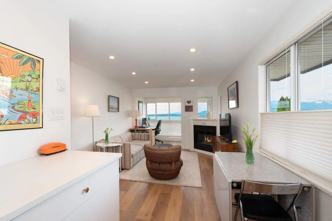Bright living room with wood floors, white walls, and recessed lighting. A sofa, desk, fireplace, and water view are visible.