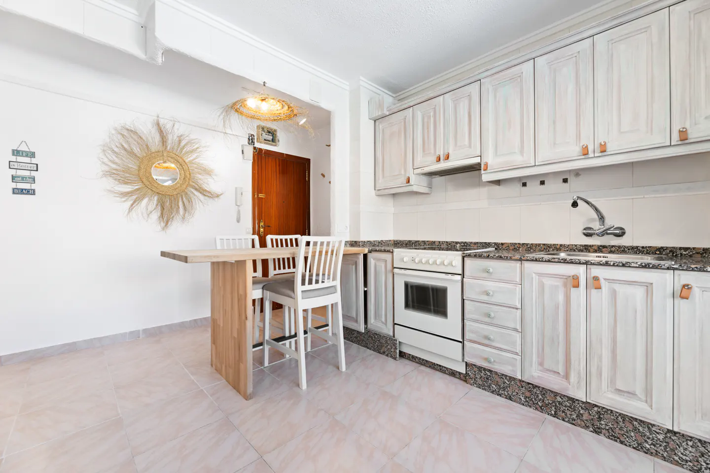 Bright kitchen with white cabinets, granite countertops, and a wooden table with two chairs. A straw sun mirror hangs on the wall.