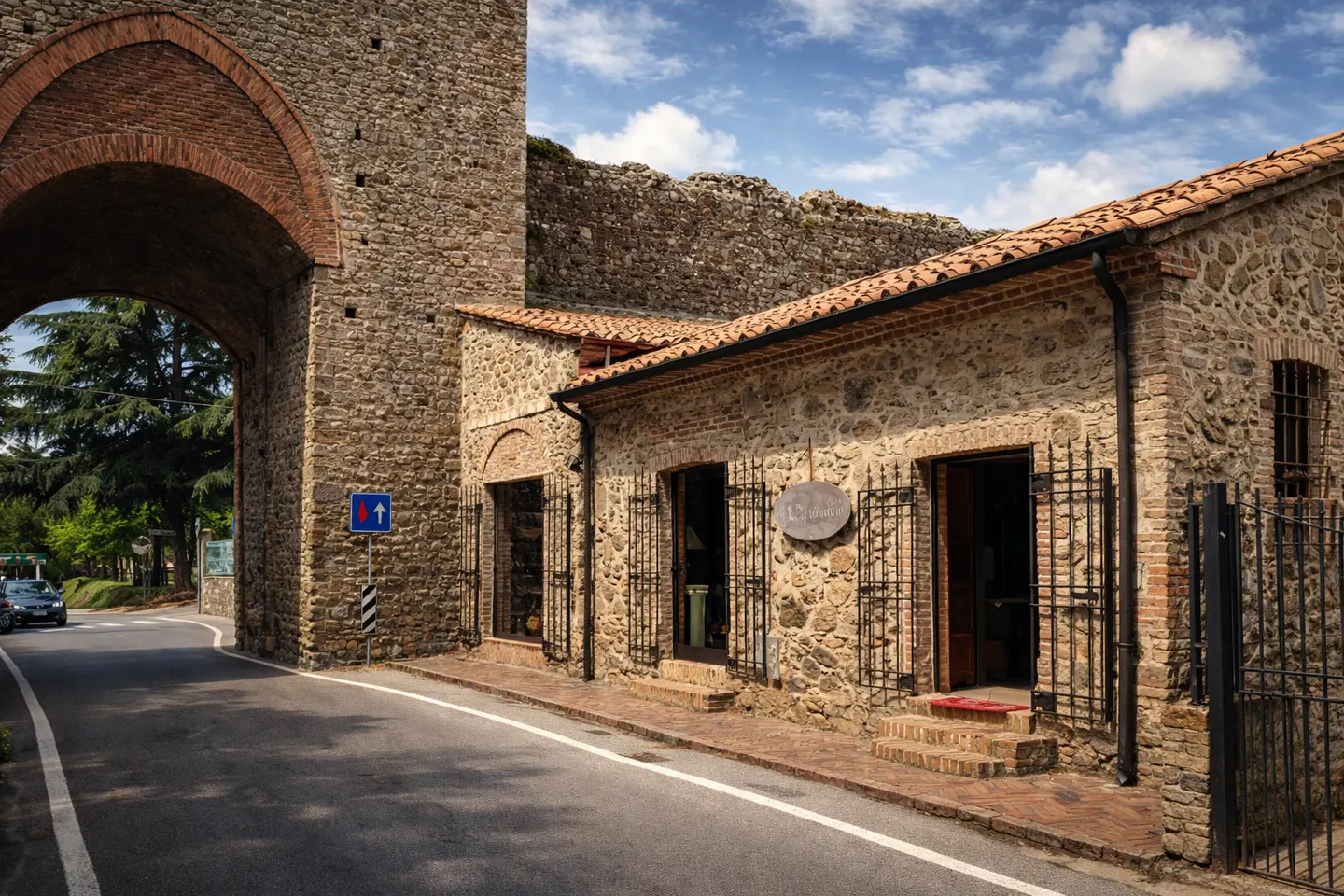 Stone building with red tile roof, seen through a stone archway on a sunny day. A road runs in front.