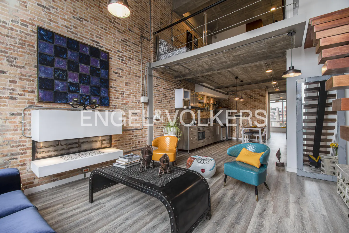 A modern loft apartment with exposed brick walls, a metal coffee table, and colorful chairs. A loft area is visible above the kitchen.