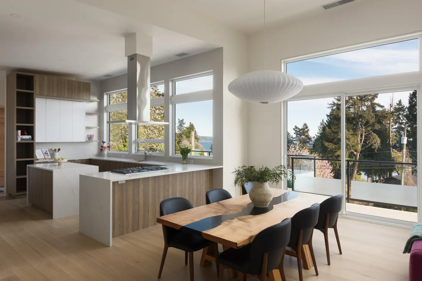 Bright, modern kitchen and dining area with wood floors. A live-edge table with black chairs sits under a white pendant light. Large windows offer a view of trees and water.
