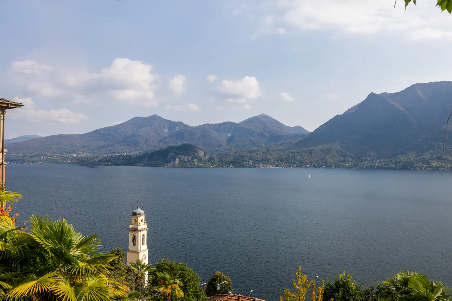 Scenic view of Lake Maggiore, Italy, with mountains, a church tower, and lush green trees under a blue sky.