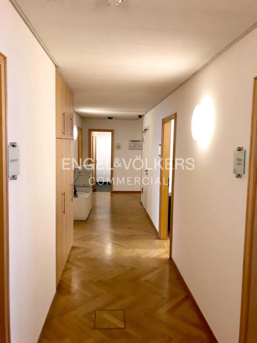 Hallway with herringbone wood floors, white walls, and wood doors. Cabinets and office equipment are on the left. Engel & Völkers logo in the center.