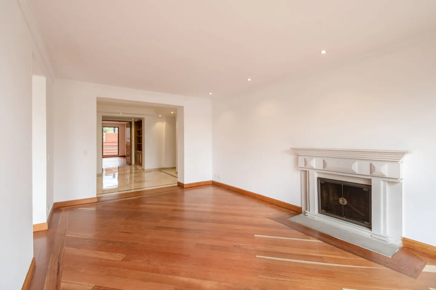 Bright, empty living room with hardwood floors, white walls, and a white fireplace. An open doorway leads to another room.