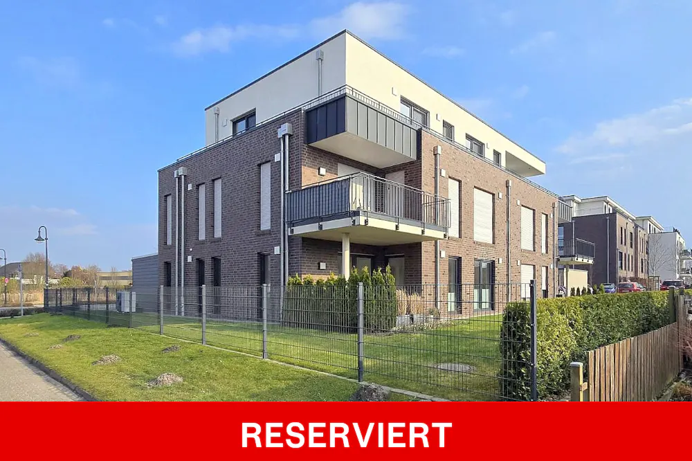 Exterior view of a modern, multi-story apartment building with brick facade and balconies, surrounded by a green lawn and metal fence. "RESERVIERT" banner.