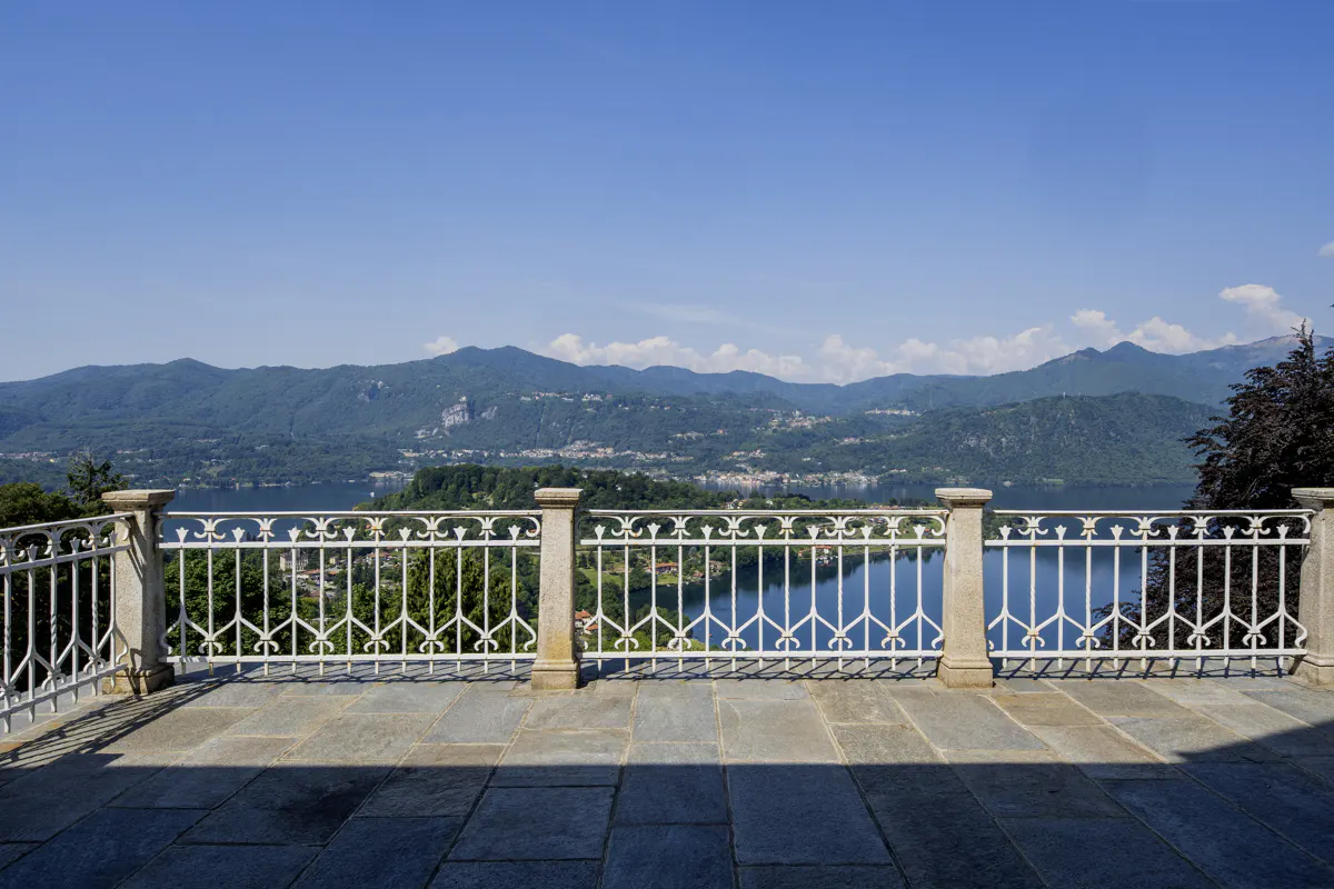 Stone terrace with a white decorative metal railing overlooking a blue lake and distant mountains under a clear blue sky.