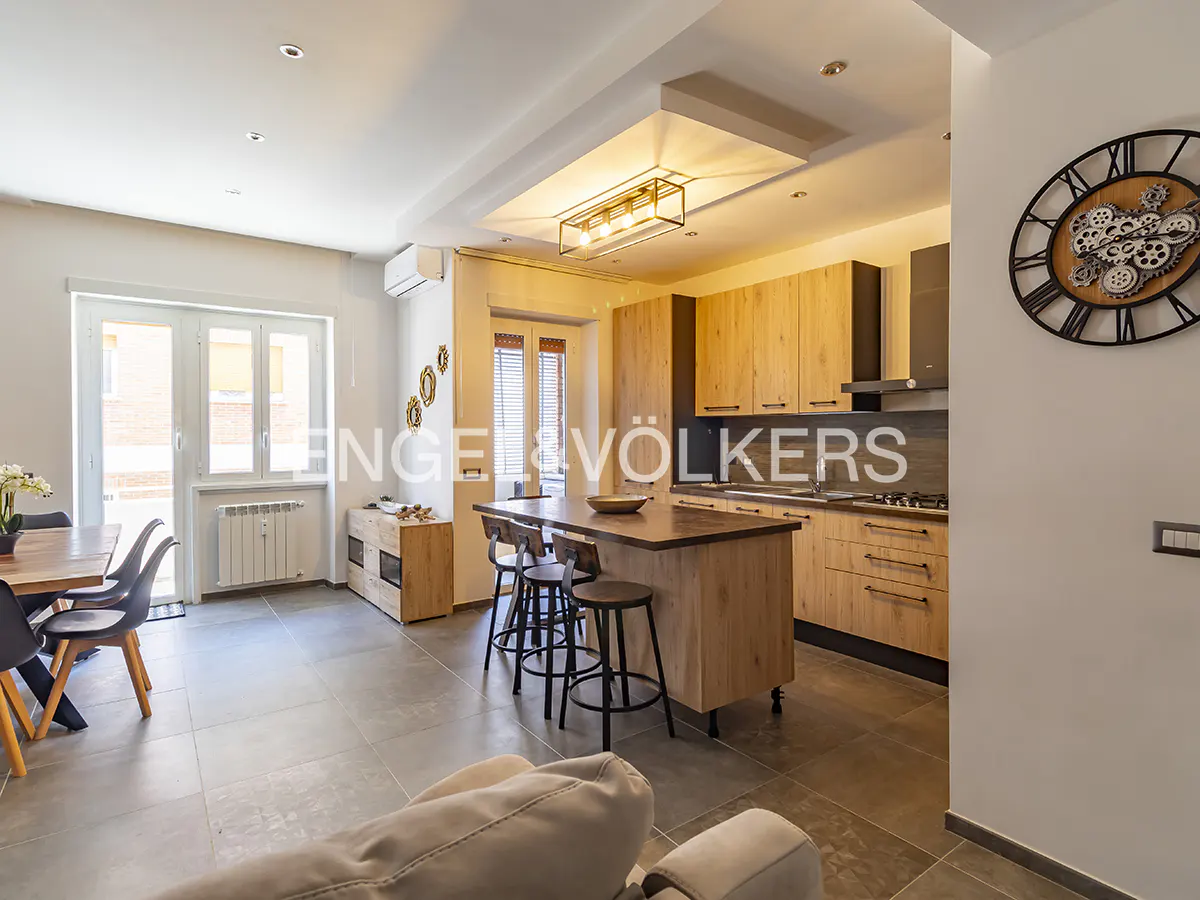 Modern open-plan kitchen with light wood cabinets, a central island with bar stools, and a dining area with gray chairs.