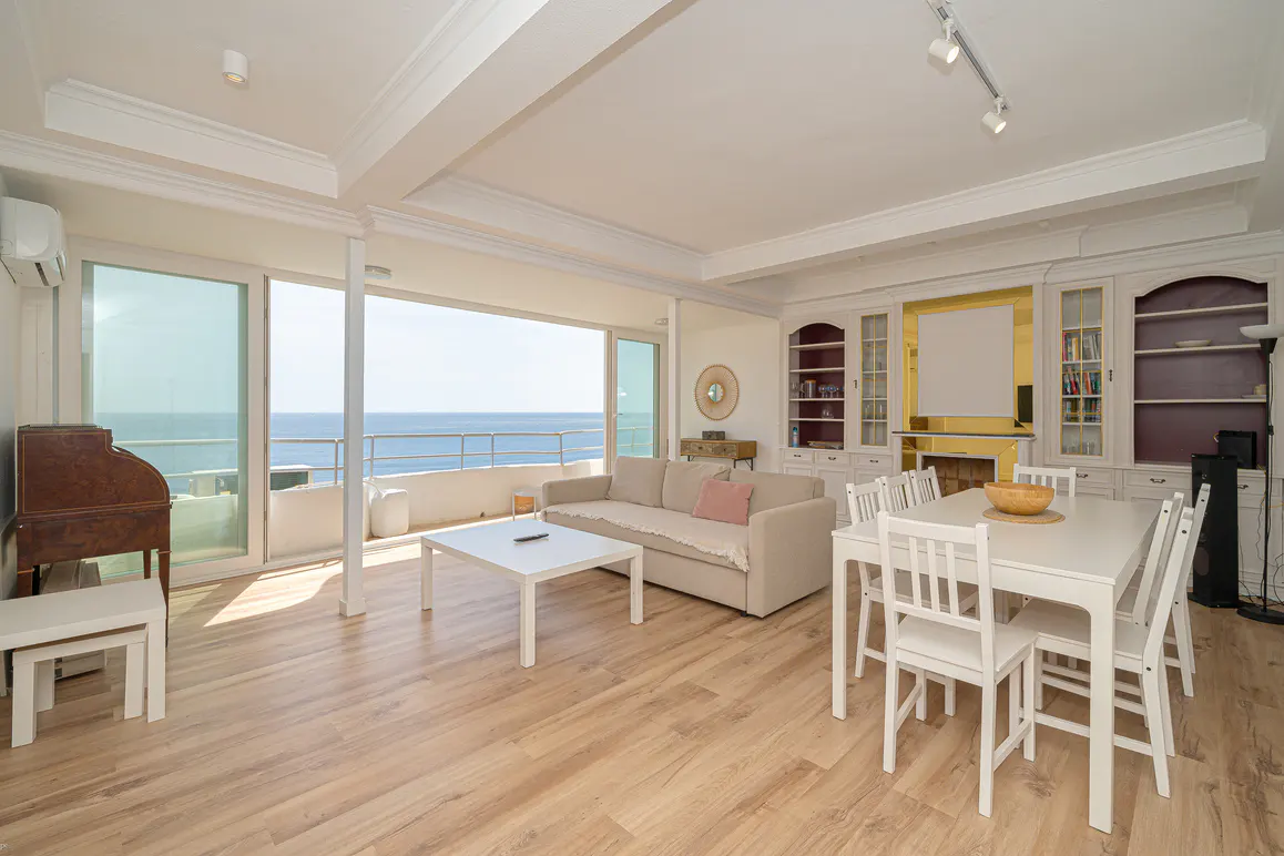 Bright living room with wood floors, white walls, and ocean view. Beige sofa, white table and chairs, and built-in bookshelves.