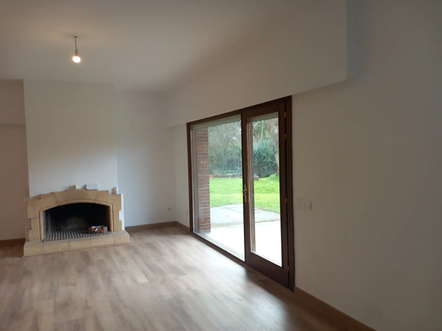 Bright living room with light wood floors, white walls, and a stone fireplace. Sliding glass doors lead to a green backyard.