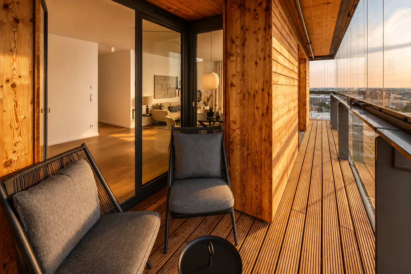 Modern balcony with wooden walls and flooring, two grey chairs, a small table, and a view of the city at sunset.