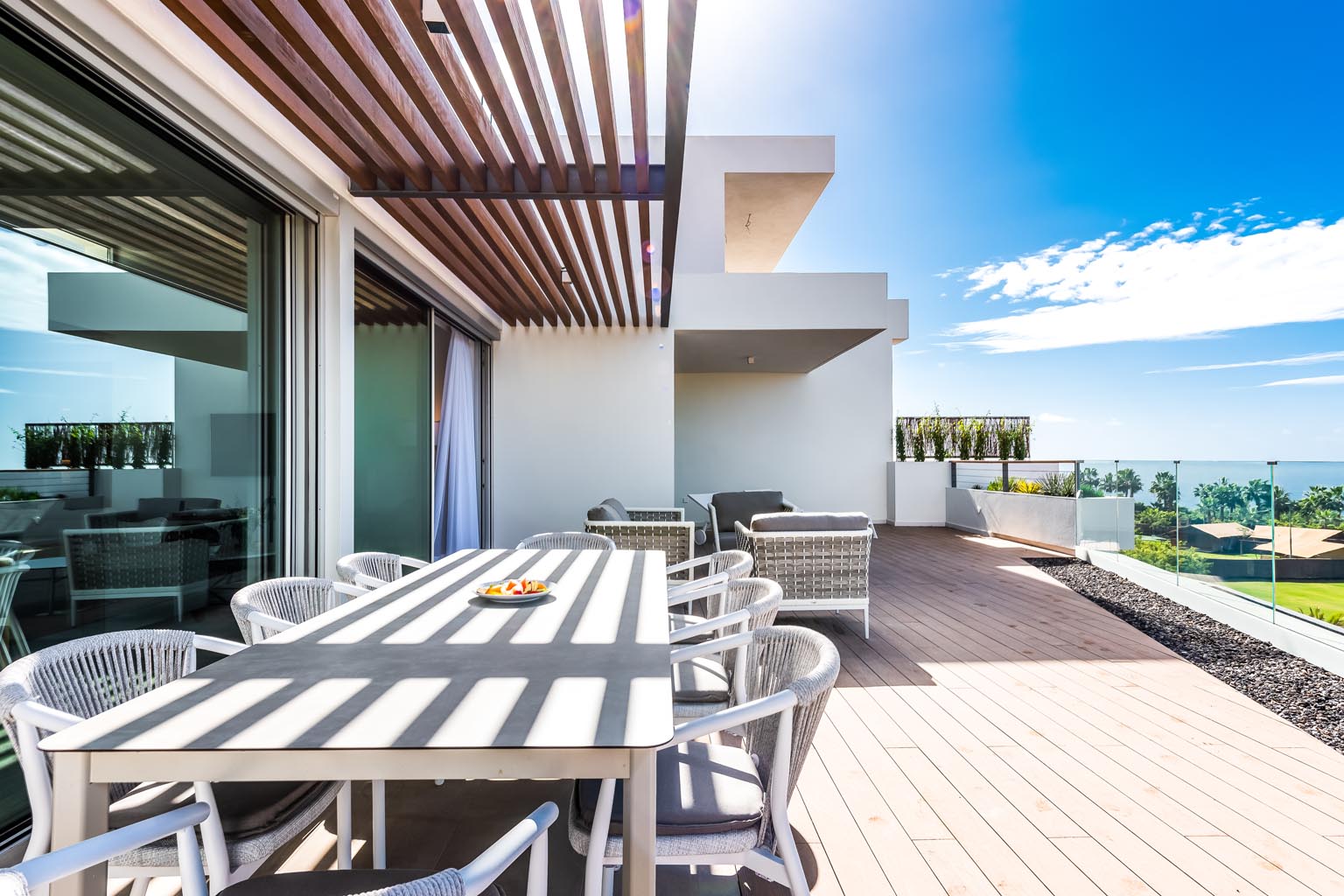 Outdoor patio with a dining table, chairs, and lounge seating on a wooden deck with an ocean view under a bright blue sky.