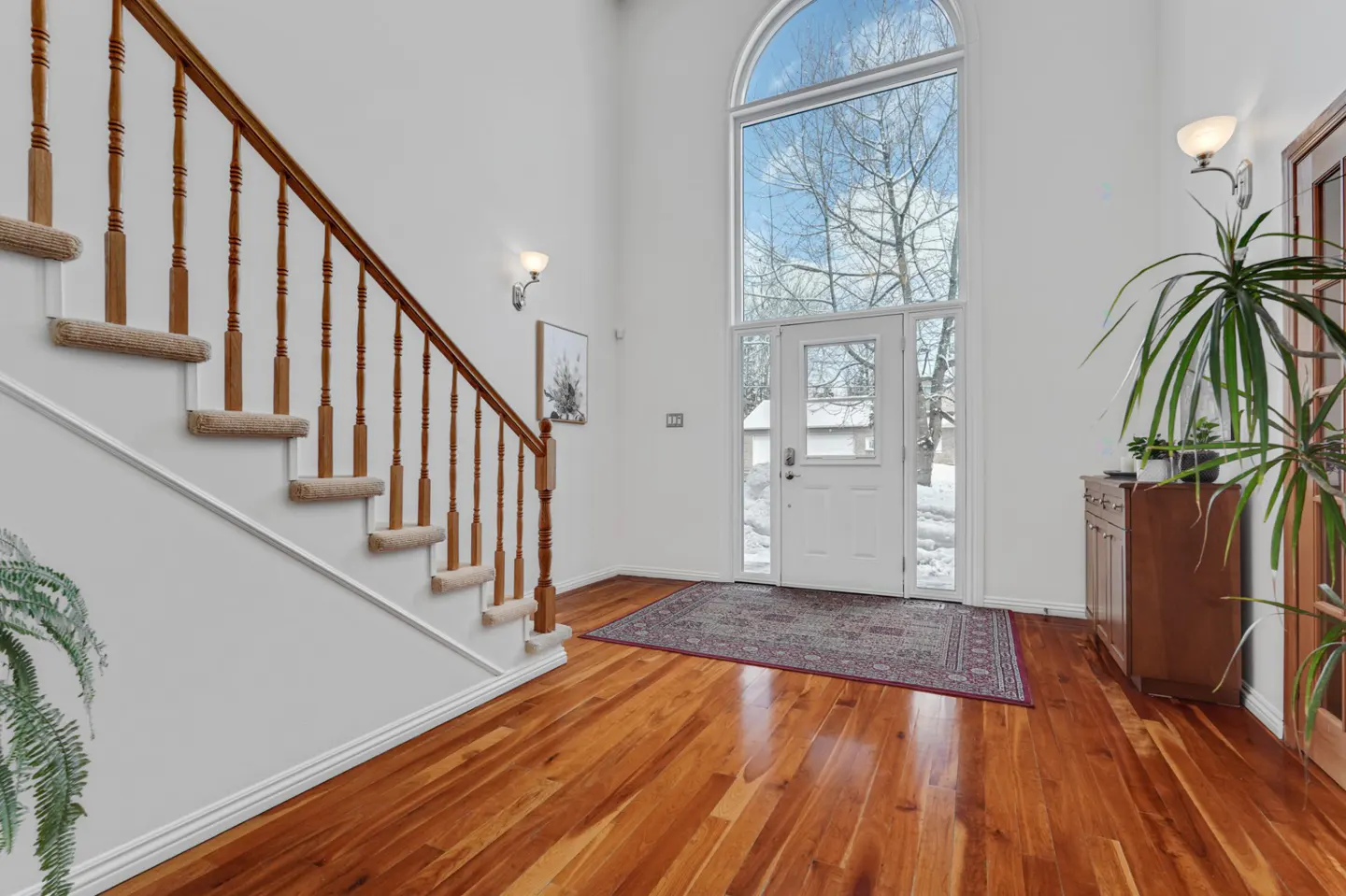 Bright foyer with hardwood floors, white walls, and a staircase. A large arched window above the front door lets in natural light.