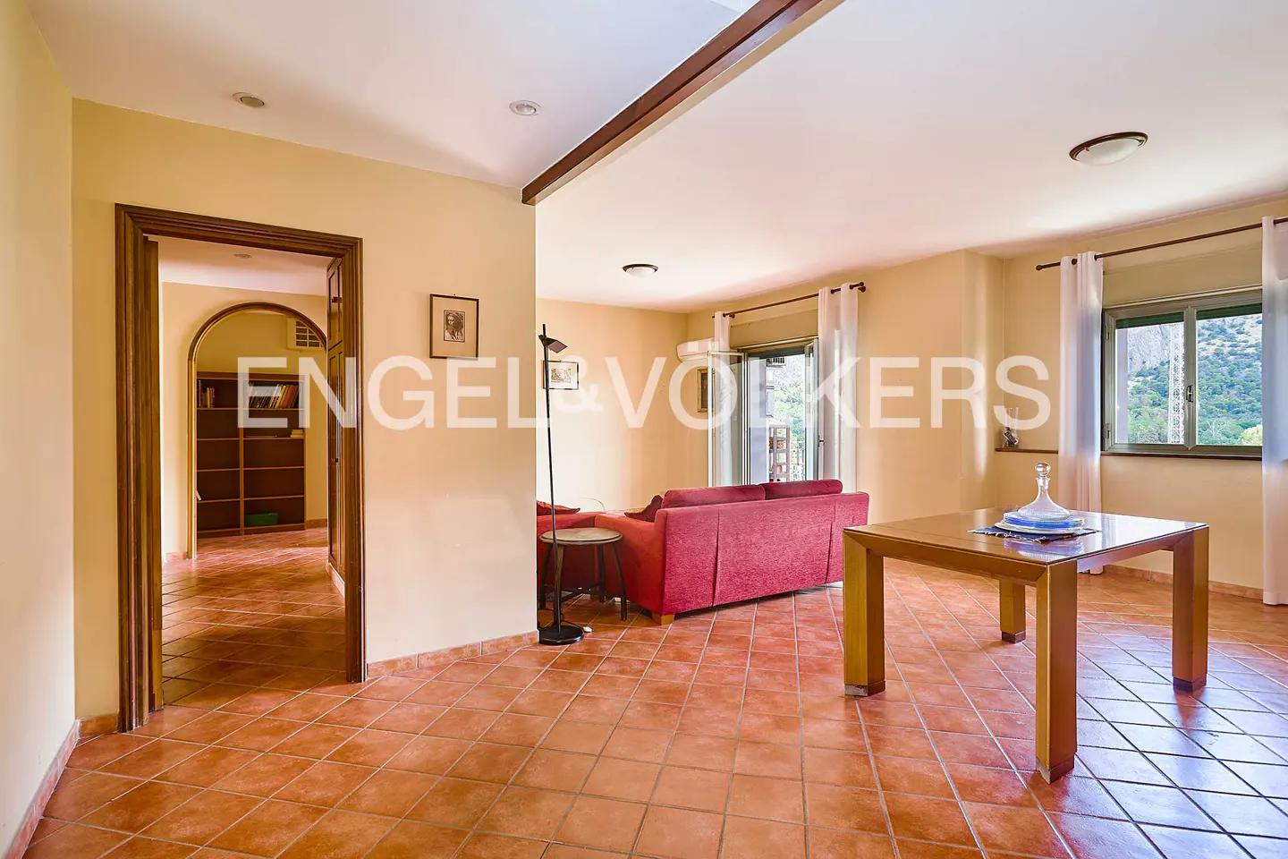 Spacious living room with terracotta tile floors, a red sofa, a wooden dining table, and a doorway leading to a library.