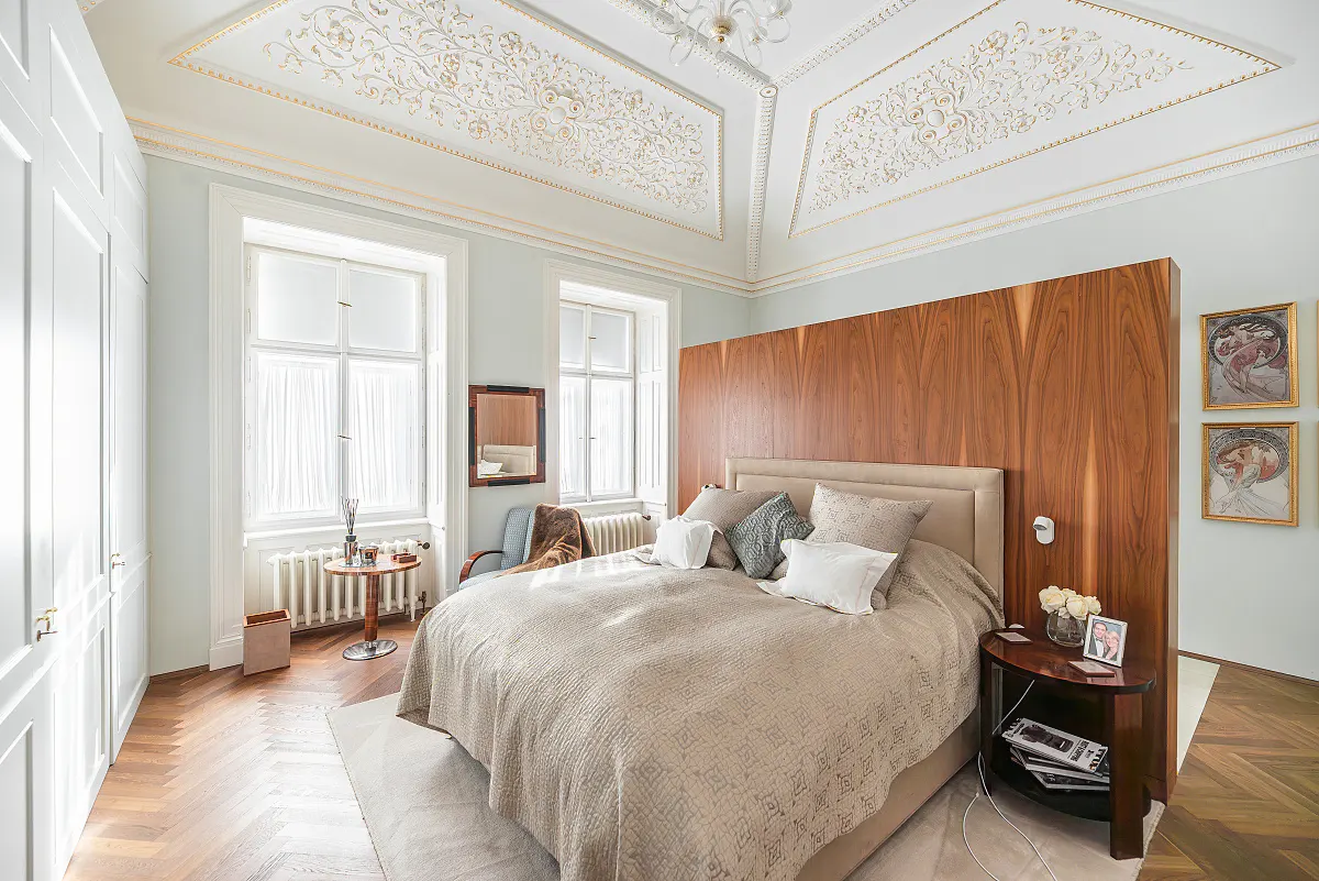 Bedroom with a large bed, wood headboard, and ornate ceiling. Two windows let in natural light. A small table sits beside the bed.
