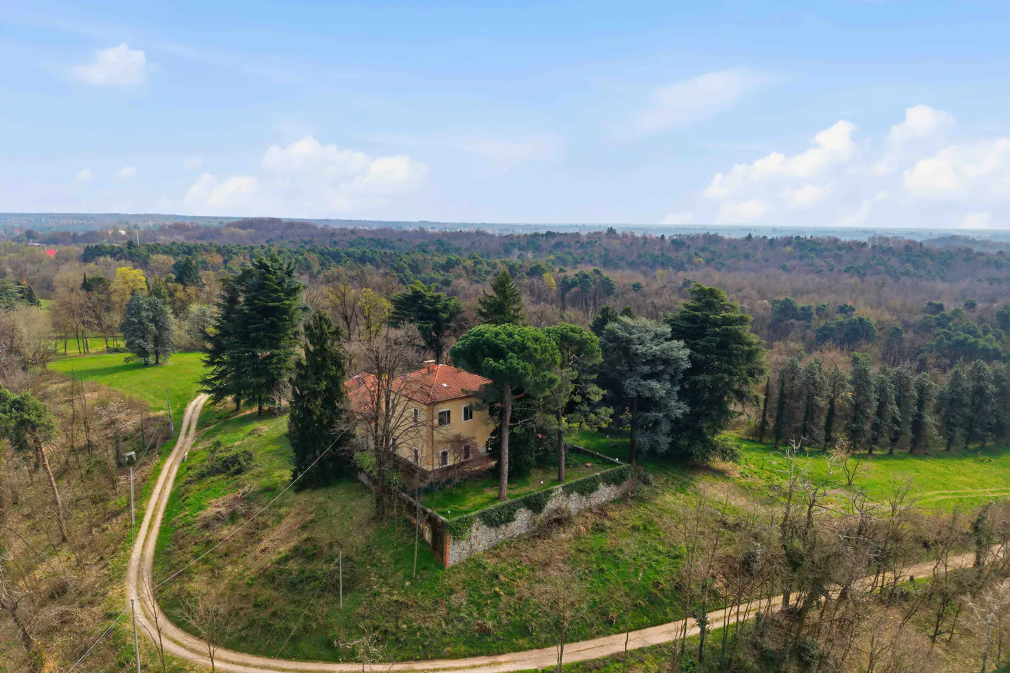 Aerial view of a yellow house with a red tile roof, surrounded by trees and a stone wall on a grassy hill.