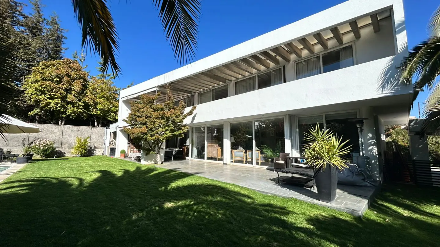 Modern white two-story house with a green lawn, patio, and trees under a blue sky.