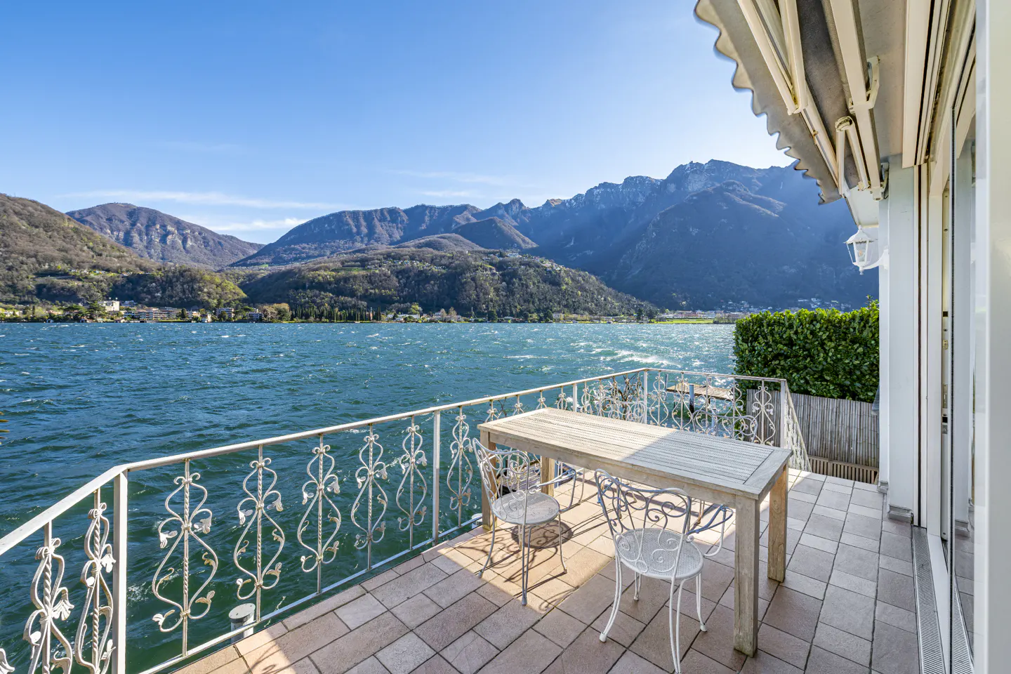 A tiled balcony with a wooden table and white chairs overlooking a blue lake and mountains under a clear sky.