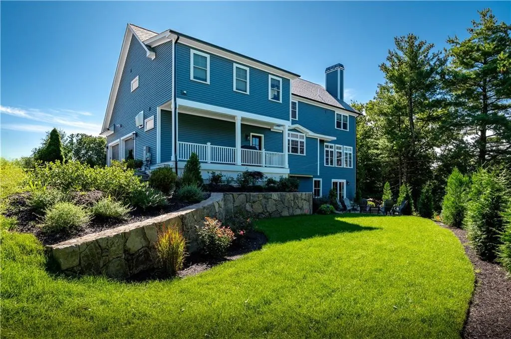 Two-story blue house with a white porch, stone retaining wall, and green lawn under a clear blue sky.
