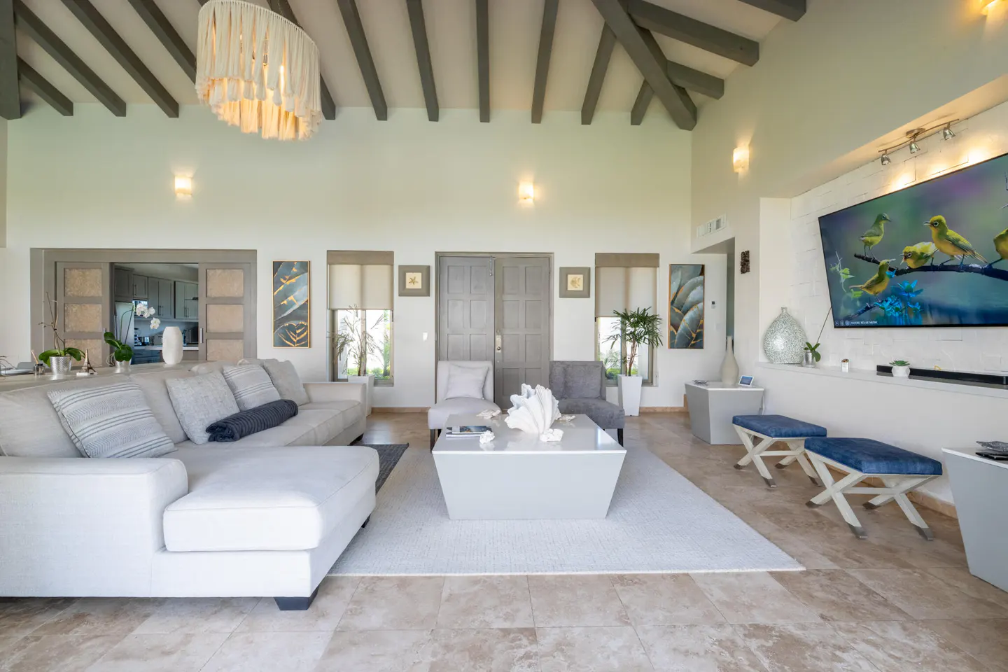 Bright living room with a white sectional sofa, gray chairs, and a large TV showing birds. The room has high ceilings with exposed beams.