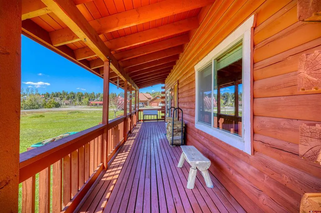Long, covered porch of a log cabin with a wooden bench, firewood, and a view of a green field and blue sky.