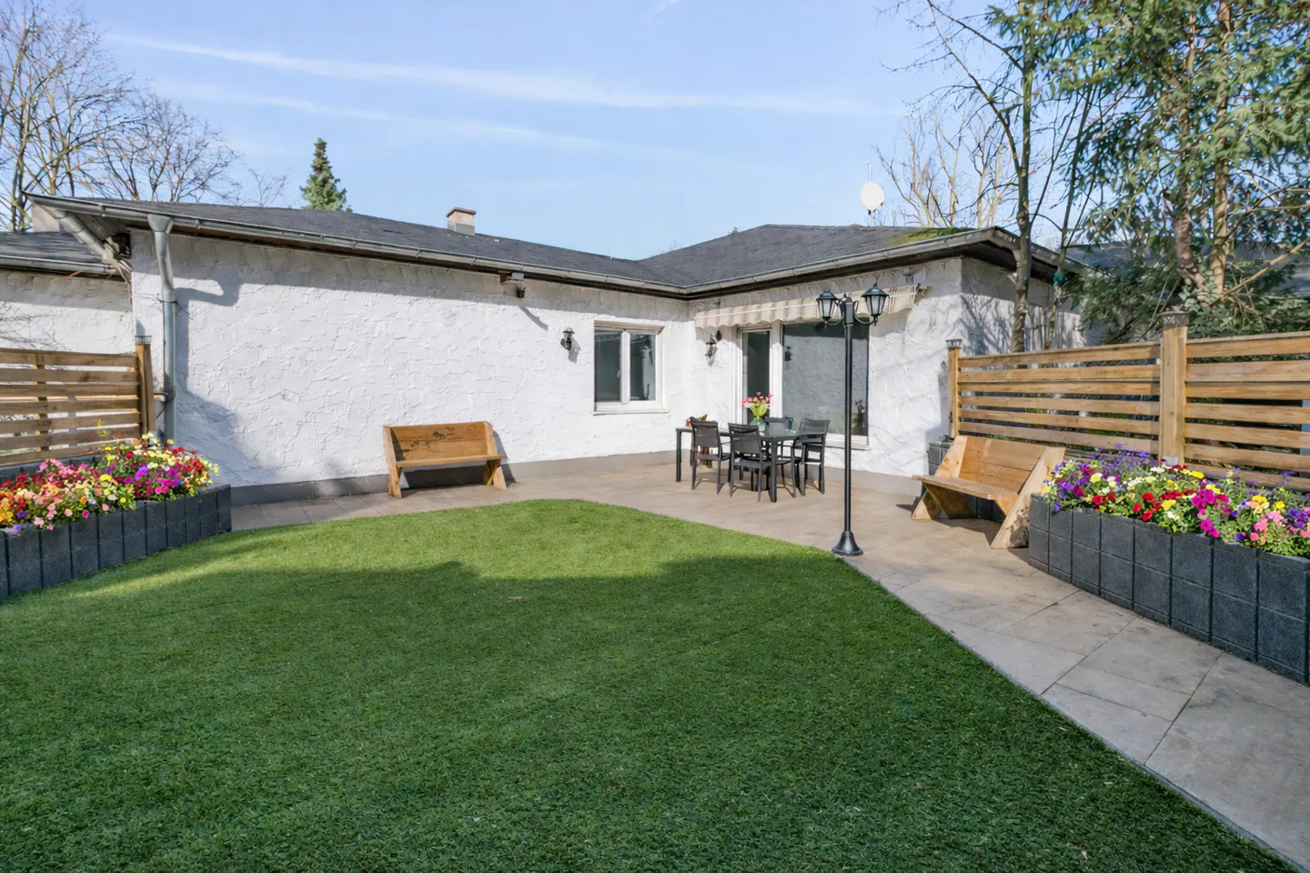 A backyard with green grass, a patio with a table and chairs, benches, and flower beds. The house is white with a dark roof.