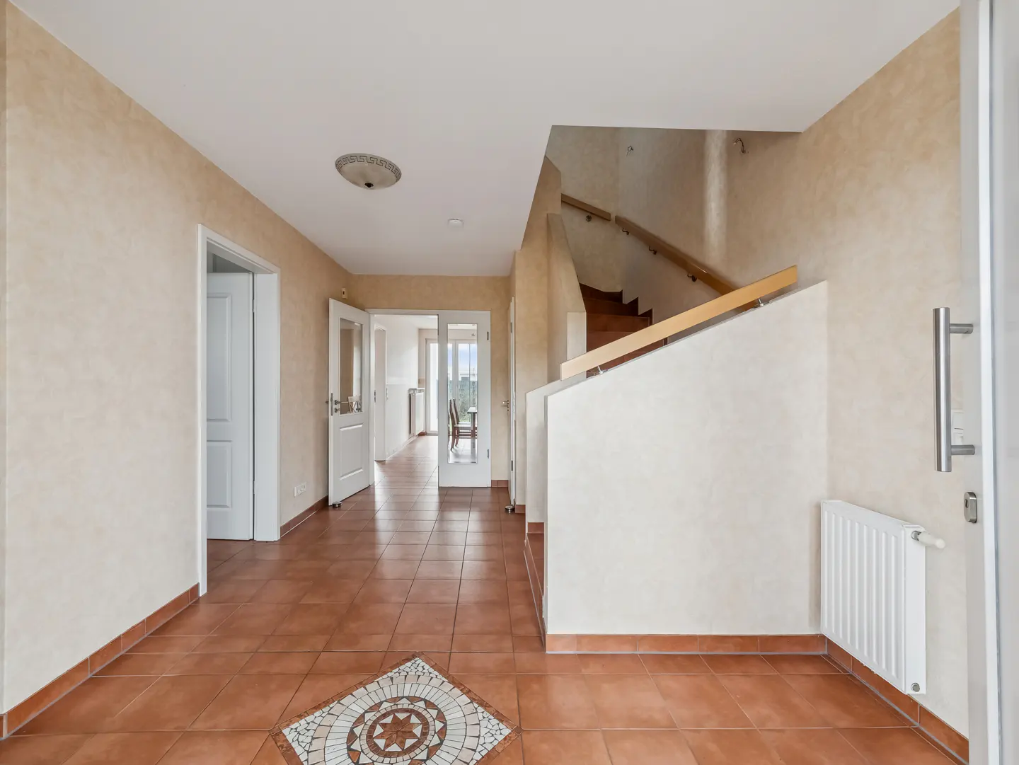 A bright foyer with beige walls, brown tile floors, and white trim. A staircase is visible on the right, and doorways lead to other rooms.