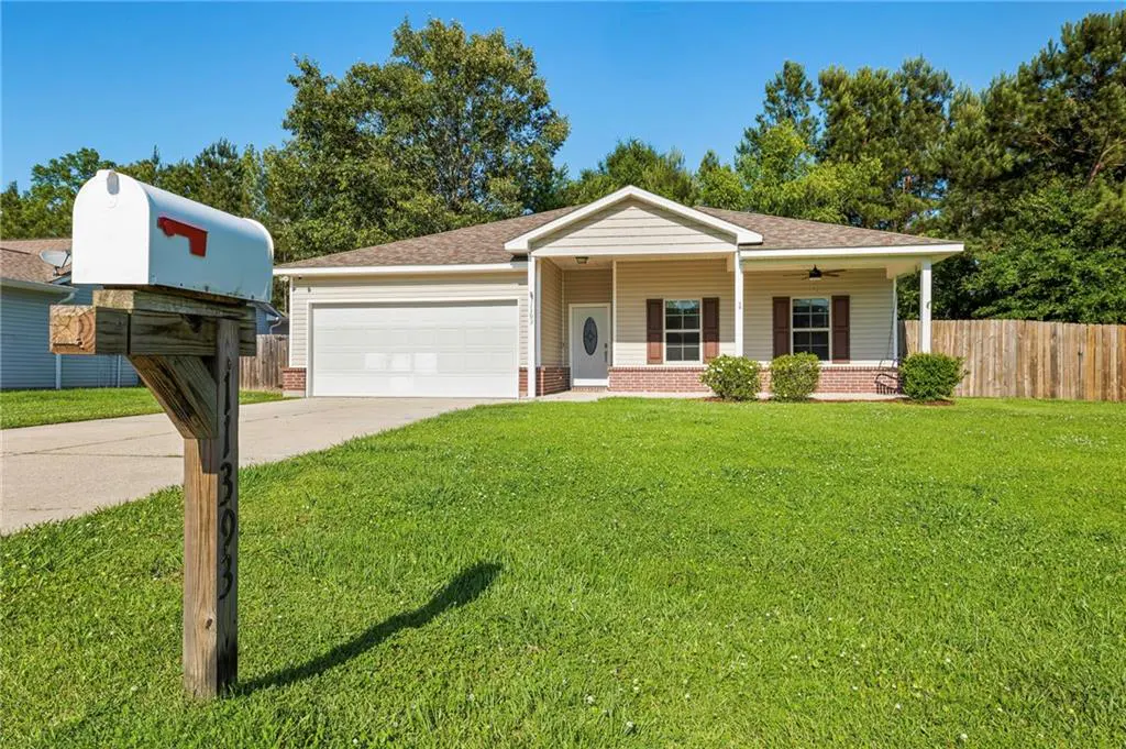 A single-story beige house with a white garage door and a green lawn on a sunny day. A white mailbox stands in the foreground.