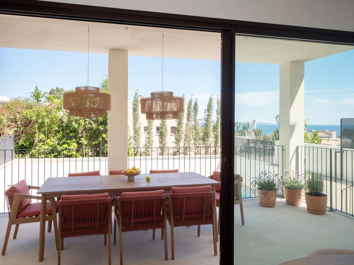 Outdoor dining area with a wooden table, red chairs, and woven pendant lights on a balcony overlooking the ocean.