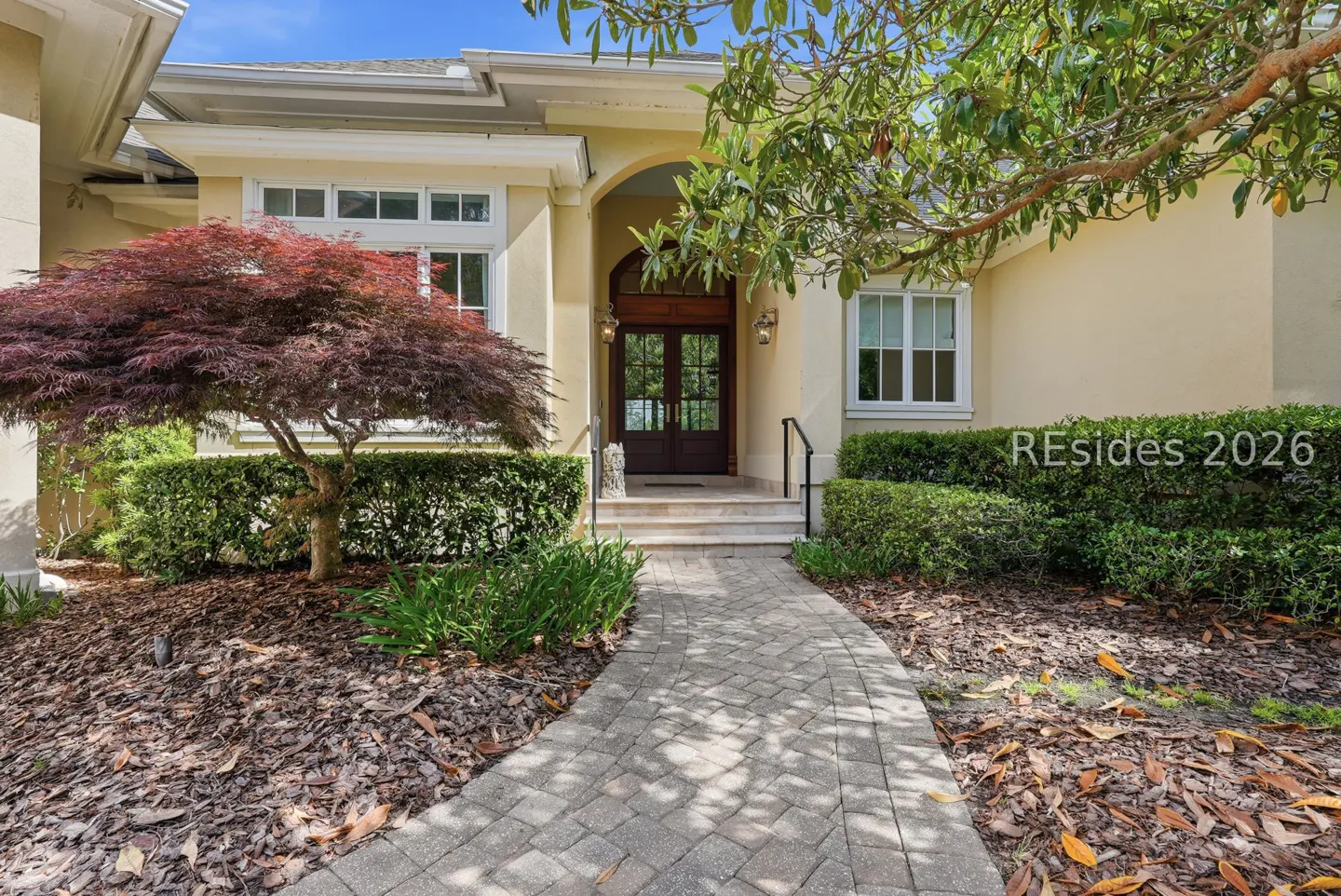 Exterior of a cream-colored home with a brick walkway leading to a dark wood double door. A red Japanese maple tree is on the left.